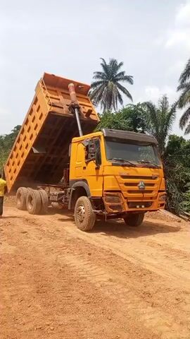 A yellow Sinotruk Howo dump truck is shown stationary on a dirt track, actively unloading material from its raised bed. The truck's hydraulic cylinder is fully extended, tilting the cargo bed upward. At least one person is partially visible near the rear of the truck. The scene is outdoors on a bright, sunny day, with a clear sky overhead. Palm trees and lush green vegetation frame the background, suggesting a tropical or subtropical environment. The ground consists of reddish-brown earth, characteristic of soil often found in regions like Aloko, Nigeria.