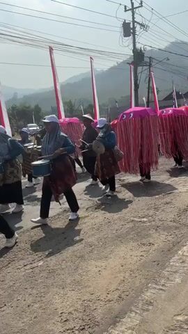 A group of people are marching in a parade outdoors. The people in the foreground are wearing headscarves and patterned skirts. One person is playing a drum. Behind them, several people are carrying large, pink, umbrella-like structures adorned with red tassels. These structures are tall and appear to be designed to resemble jellyfish. Tall, red and white striped flags are also visible in the background. The parade is taking place on a dirt road, with hills and trees in the distance. The sky is clear and sunny.
