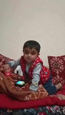 A young child lies on a red patterned bed, propped up on their arms. They are wearing a blue shirt, jeans, and a red vest with floral patterns. A smartphone is on the bed near them, and they are holding a green toy car in their right hand. A brown blanket is partially covering the bed. There are two red patterned pillows behind the child. The wall behind the bed is plain white. The child looks directly at the camera. The child then looks down at their lap.
