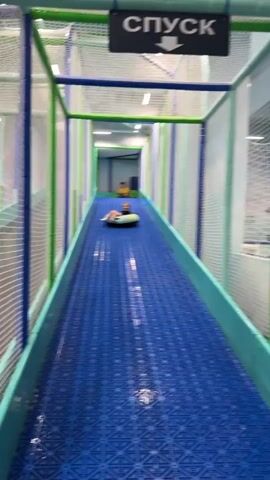 A young child is riding down a bright blue, textured slide inside an indoor play area. The child is sitting on a round, yellow inflatable tube, holding their arms up in the air with a joyful expression. They are wearing a t-shirt and shorts. The slide is enclosed by a structure with green, blue, and white mesh netting, creating a tunnel-like appearance. In the background, another child and an adult can be seen further up the slide.

Above the slide, a black sign with white Cyrillic text is visible. The text reads "СПУСК," which translates to "DESCENT" or "SLIDE" in English, and features a white arrow pointing downwards. The overall scene suggests a fun and active play environment, likely indoors due to the lighting and enclosed structure. The colors are vibrant, contributing to a playful atmosphere. The child's body language and facial expression clearly indicate excitement and enjoyment.