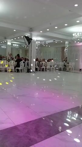 The video shows a wide shot of a large, well-lit indoor venue, likely a banquet hall or reception area. The space has a high, white ceiling with multiple chandeliers and recessed lighting fixtures. The floor is polished white marble with dark veining, reflecting the light and appearing wet or illuminated with colored lights, primarily pink and purple.

In the background, numerous round tables are set with white tablecloths and chairs, indicating a gathering or event. People are seated at these tables, engaged in conversation or observing something. Several pillars are visible, decorated with white fabric and some with what appear to be hanging decorations or lights.

A prominent object in the middle ground is a large, parabolic light-reflector umbrella mounted on a stand, aimed towards the ceiling or a central area. There are also speakers and audio equipment visible, suggesting music or a performance.

In the foreground, slightly to the right, a man in a dark suit is standing and appears to be playing a violin. He is facing away from the camera and towards the seated guests. Another person, possibly a woman in a white dress, is standing near the man with the violin, and there might be a floral arrangement or decoration between them. The camera perspective is low, looking across the dance floor towards the seated guests and the performer. The overall atmosphere appears to be celebratory, possibly a wedding reception or formal dinner.