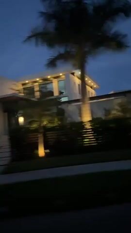 This is a blurry, shaky, handheld video taken at dusk or night from a moving vehicle, likely a car. The scene shows a large, modern house with contemporary architecture, featuring a flat roof, large windows, and exterior lighting that illuminates the facade and landscaping. Several palm trees are visible, framing the house and contributing to a tropical or subtropical environment. The lighting suggests it is either sunrise or sunset, or that the house's exterior lights are on. The motion blur indicates the vehicle is in motion, possibly driving past the property.