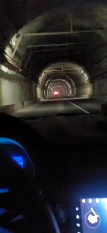 A point of view shot from inside a vehicle driving through a tunnel. The tunnel is dimly lit with overhead lights and the perspective draws the viewer's eye toward the red taillights of a vehicle ahead. The dashboard of the vehicle is partially visible in the foreground, with a blue digital display on the left. The tunnel walls are rough and appear to be made of concrete, with pipes and cables running along the ceiling. The road is paved and has a white line marking the lane. The overall impression is one of movement through a dark, confined space.