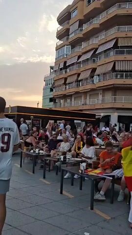 The video shows a street scene with a crowd of people sitting at outdoor tables of restaurants or bars. Many of the people are wearing red and yellow, the colors of the Spanish flag, and some are holding Spanish flags. There is a tall building with balconies in the background, and palm trees are visible. Some people are walking down the street, while others are seated and engaged in conversation. One person is carrying a Spanish flag draped over their shoulders. The atmosphere appears to be festive. The video transitions to show more of the street with people walking and palm trees. Some individuals are seen holding Spanish flags.
