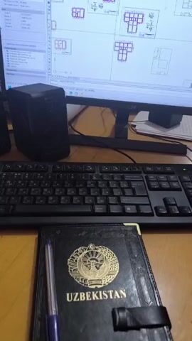 The video shows a desk with a black keyboard, a black notebook with a golden emblem, and a silver pen. In the background, a computer monitor displays a blueprint on a white background. A black speaker is positioned to the left of the monitor. The camera moves slightly to show the full computer screen. The blueprint contains multiple outlines of structures, some highlighted in red and others in blue, along with various technical details and annotations. A webcam is affixed to the top of the monitor. An upbeat musical audio track is playing in the background.