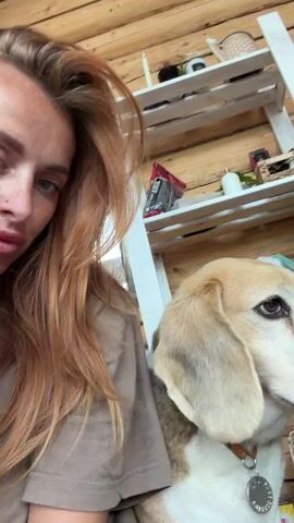A woman with long, light brown hair is taking a selfie video. She is sitting next to a beagle. The woman is looking down at her phone, and the dog is looking to the side. The background is a wooden wall with shelves and various objects on them.