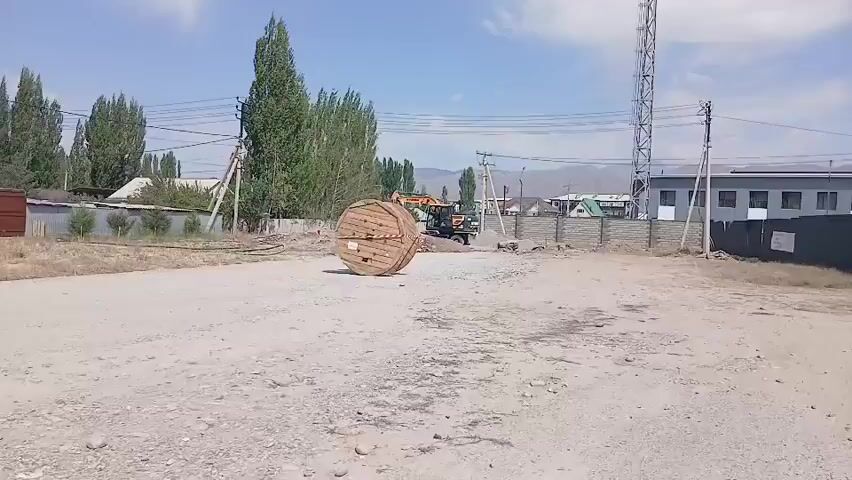 The video shows a construction scene on a sunny day. In the foreground, there is a large wooden spool lying on a dirt road. Behind the spool, an excavator is seen operating. Further in the background, there are trees, power lines, and buildings. The overall environment appears to be an industrial or construction area with some signs of ongoing work.