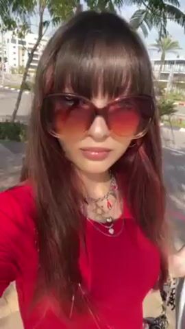 A woman wearing sunglasses, a red shirt, and blue pants is walking outside. She sticks her tongue out at the camera. A sidewalk and a street with cars can be seen in the background.