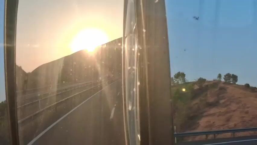 The video shows a view through a vehicle's side mirror as the sun sets. The mirror reflects a highway with a guardrail and a lone car passing by. The sun is low on the horizon, casting a bright glare over the scene, and a hilly landscape with sparse vegetation is visible on either side of the road. The sky is clear and blue, with a few specs of dust or dirt visible on the mirror's surface.