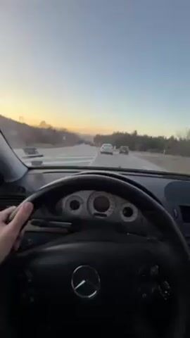 The video shows a view from the driver's seat of a Mercedes-Benz, driving on a highway during sunset. The driver's hand is visible on the steering wheel. Other cars are also traveling on the highway. The sky is clear, with colors ranging from orange near the horizon to light blue above. The roadside is lined with trees.