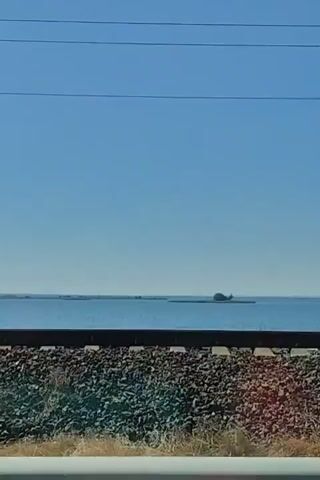 The video displays a body of water with a clear blue sky above. In the foreground, there are railroad tracks and ballast. In the distance, there is a small structure or boat on the water. Several utility lines are visible stretching across the sky. The camera appears to be moving, likely from a vehicle, as indicated by the slight motion blur and the presence of a railing in the bottom of the frame. The scene is brightly lit, suggesting daylight.