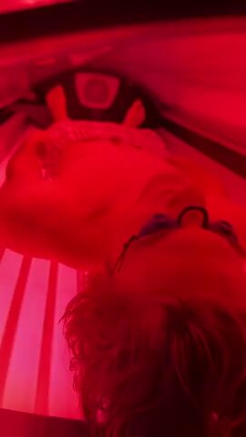 The video shows a person lying down inside a tanning bed, which is illuminated with red light. The person is wearing goggles to protect their eyes. Their body is partially visible, and they appear to be relaxed. The tanning bed is a common piece of equipment used for artificial tanning. The environment is dark except for the bright red light emanating from the tanning bed.