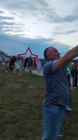 The video shows an outdoor gathering of people under a cloudy sky. In the foreground, a man in a blue patterned shirt and jeans is holding up a smartphone, seemingly taking a picture or recording video. In the background, there are other people, some standing and some walking, and a large red star decoration. The overall atmosphere appears casual, possibly at an event or festival.