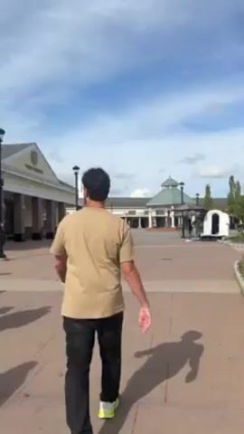 A person walks down a brick pathway toward a shopping center. The person is wearing a tan shirt and dark pants. To the person's right is a small landscaped area. Beyond that is a Jimmy Choo store. Beyond the Jimmy Choo store are other stores. In the distance is a building with an octagonal roof. The sky is blue with some clouds. A white cart is parked near the brick pathway. A trash receptacle is to the right of the cart. The person points to their left.