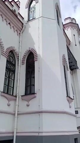 The video shows a close-up, low-angle view of a white building with pink trim. The building features a tall, slender tower with arched windows and decorative crenellations at the top. Another smaller tower is visible to the right, also with similar architectural details. The building's facade has several arched windows with dark frames, some of which appear to be stained glass. A white downspout runs along the side of the building. The lower part of the building is painted gray. The ground in front of the building is covered with gravel. The sky, partially visible at the top, is cloudy with patches of blue. The camera pans slightly upwards as it moves along the side of the building.