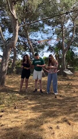 Three individuals stand in a grassy, pine-cone strewn area dotted with tall trees. A woman on the left, sporting a black tank top and black biker shorts, appears barefoot. In the middle stands a person in a green Adidas t-shirt and patterned shorts, wearing shoes. To the right, a woman in a floral shirt and wide-legged blue pants wears sunglasses and white shoes. Each person holds a yellow paper in their hands. They appear to be reading, and possibly reciting or acting out a script, as evidenced by their expressions and the way they hold the papers. Behind them, a hammock hangs between the trees. The atmosphere is sunny and relaxed, suggesting an outdoor activity or gathering.