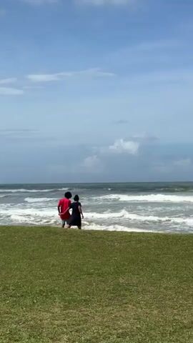 Two young children, one in a red shirt and the other in a dark shirt, stand on a grassy area facing a choppy ocean under a cloudy blue sky. They appear to be holding hands or walking together. The waves are breaking near the shore. The scene is captured from behind the children.
