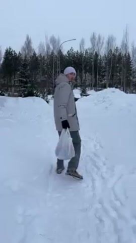 A man wearing a white beanie, light grey coat, grey pants, and black gloves walks away from the camera in a snowy landscape. He carries a plastic shopping bag in his right hand. The ground is covered in snow, and footprints mark the path he's walking. Behind him are trees, and a building is visible in the distance. The sky is overcast.
