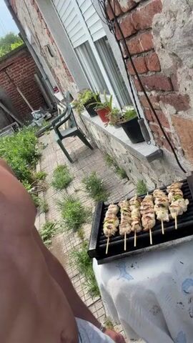 A shirtless man stands outside on a paved patio and grins into the camera, turning to show a grill with seven skewers of chicken and green peppers cooking on it. The grill is on a table with a white tablecloth. A white-shuttered window with potted plants on the sill is visible behind the grill, and a brick wall to the right.