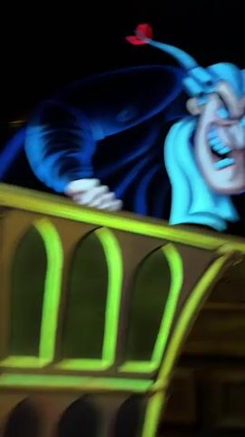 A blurred, dimly lit video shows the animated figure of a person with light blue hair and a pale face, wearing a light blue outfit and dark blue cape. The figure has a wide, toothy grin and appears to be pointing with its right hand. The background is dark with hints of a wall and what might be a painting or tapestry. The video then transitions to a dark environment with glowing yellow and blue lights, possibly representing a set or stage. The final frames show a dark tunnel with two red lights and a bright white light in the distance, resembling the front of a vehicle.