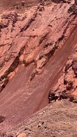 A video shows a close-up view of a steep, rocky mountainside with distinct geological layers. The terrain is predominantly red and brown, with textured rock formations and loose scree. The camera pans slowly across the landscape, highlighting the erosion patterns and the rugged, natural environment. There are no people or objects visible in the video, only the geological formations of the mountainside.