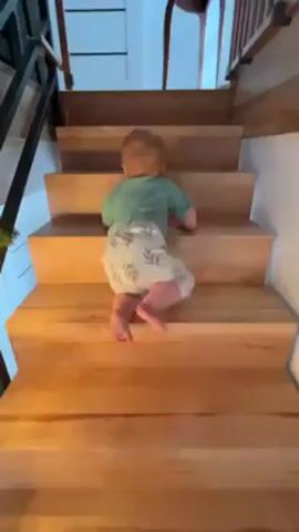 A baby wearing a green shirt and pants with a leafy pattern is climbing up a set of wooden stairs. The stairs have a light wood finish. There is a black metal railing visible on the left side of the frame.  A white paneled door can be seen next to the stairs. The camera angle is low, looking up at the baby.
