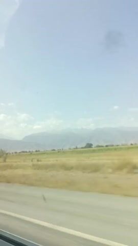 The video shows a landscape from a moving vehicle. The sky is blue with some scattered clouds. In the middle ground, there is a field with dry grass and a line of trees. In the background, there are mountains. The foreground shows a road or pavement, also blurred due to the motion. The overall impression is of a rural, possibly arid or semi-arid environment under a clear sky.