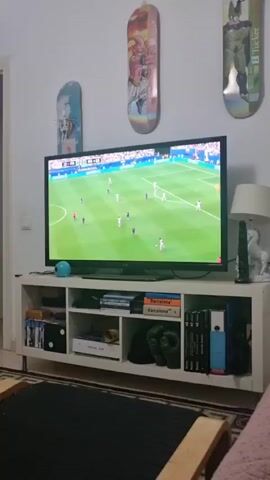 The video shows a television screen displaying a soccer game. A player in a white jersey with the number 25 on the back is dribbling a soccer ball on a green field. The score on the screen shows FRANCE 0, ENGLAND 2. The game is being played in a stadium with a crowd in the background. The television is sitting on a shelf with books and other items.
