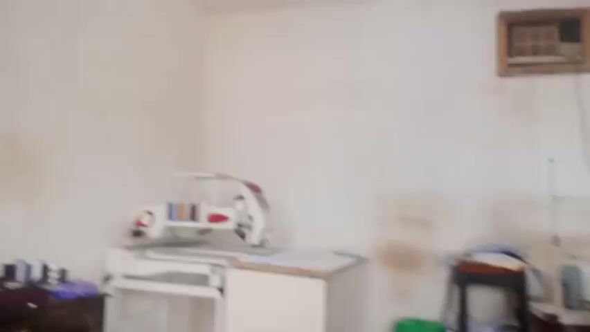 This media file is a short video capturing a glimpse of a sewing workshop. The primary subject is a man, presumably a tailor, seated at a sewing machine. He appears focused on his work, with a measuring tape draped around the machine and his neck. Several spools of thread are visible on the machine and a stand, suggesting ongoing sewing operations.

The setting is an interior space, likely a workshop or tailor's shop, with plain, light-colored walls. There are multiple sewing machines present, including the one the man is using and another larger machine on a table to the left, which also appears to have a design or pattern on it. Other objects in the scene include a stool with some folded clothes on it, a small green stool, a table with papers, and what looks like a small appliance or ventilation unit mounted high on the wall. A dark wooden cabinet or dresser is partially visible on the far left.

The lighting suggests it could be daytime, but the exact time of day is not discernible. There are no clear indications of weather conditions. The overall atmosphere seems to be one of quiet work and industry. The man's expression is neutral and concentrated. No discernible text is visible in the video.