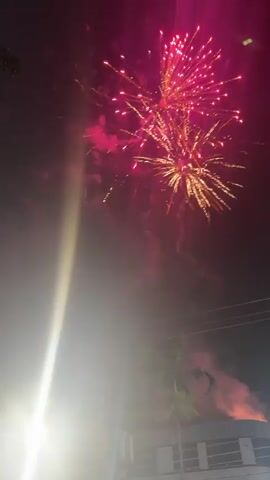 The video shows a nighttime scene with fireworks being set off. The sky is dark, and sparks and light trails from the fireworks illuminate the darkness. There is a bright light source in the lower left corner of the frame, which could be a street light or a camera flash. In the background, the silhouettes of trees and buildings can be seen, suggesting an outdoor urban or suburban setting. Some of the fireworks create bursts of light and smoke, while others leave trails of sparks as they ascend or descend. The overall atmosphere is festive, indicative of a celebration or event.