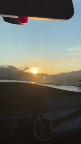From the interior of a car, the camera captures a view of a sunrise over a slightly hilly landscape. The sun is low on the horizon, partially obscured by trees, casting a bright glare. The sky transitions from a light blue near the horizon to a deeper blue higher up, with a few thin clouds. The car's dashboard is visible in the foreground, along with the rearview mirror. The windshield has some minor smudges. The car appears to be moving, as the landscape slowly shifts.