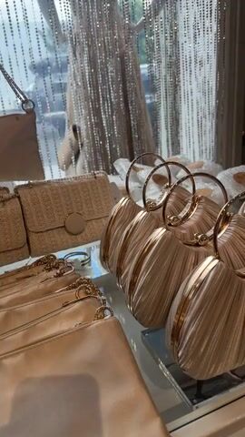 The camera pans slowly across a collection of gold and cream-colored purses displayed on a glass-topped white table. The purses, which are arranged in rows, have round gold handles and textured pleated fabric. In the background, dresses hang on a rack, and beaded curtains and a display cabinet add depth to the shot. The scene appears to be inside a boutique or high-end retail store.