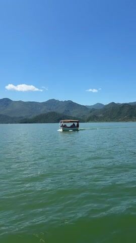 A white boat with a brown canopy carrying several passengers travels from left to right across a wide body of green water. The boat leaves a white wake behind it. In the background, a continuous range of green, tree-covered mountains stretches under a clear blue sky with a few wispy white clouds. The water shows slight ripples.