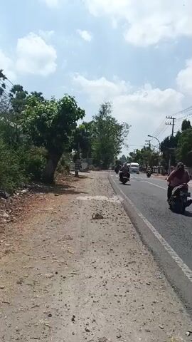 The video shows a road scene from a sidewalk. There are trees and vegetation on the left side of the road. The road itself is paved with asphalt and has white lines marking the lanes. Several motorcycles are visible on the road, as well as at least two white vans. The sky is partly cloudy with a bright sun, suggesting it is daytime. The scene appears to be in a semi-rural or suburban area, with power lines visible overhead.