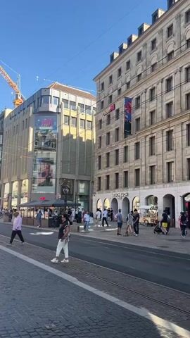 This video captures a bright, sunny day in a European city, likely Geneva, Switzerland. The scene is a bustling street with a tram line running through it. On the left, a tall building with a glass facade displays advertisements, and a construction crane looms in the background, suggesting ongoing development. Several people are walking on the cobblestone sidewalk, some crossing the street.

To the right, a modern white and red tram is in motion, its side displaying the "tpg" logo, indicating it's part of the Geneva public transport system. Adjacent to the tram tracks is another building with arched doorways and shops, one of which appears to be "AESCHBACH 1890." Further down the street, more people are visible, engaged in various activities like shopping or commuting. The overall atmosphere is one of a vibrant urban environment during daylight hours. The clear blue sky and the bright sunlight suggest pleasant weather. The presence of fashion stores like "H&M" further hints at a commercial district. There are no overt displays of emotion, but the scene generally conveys a sense of everyday activity and normalcy.