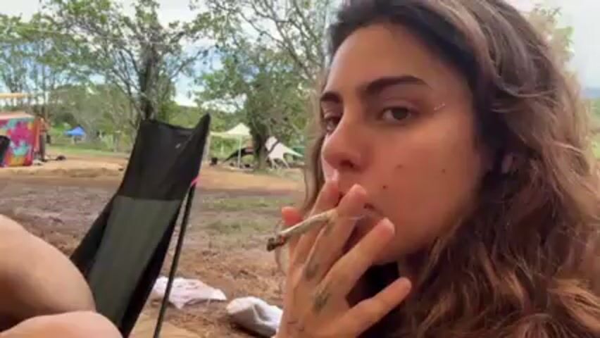 A woman with long curly hair is sitting in a black folding chair and smoking a cigarette. She has a tattoo on her hand and sparkles on her cheek. She exhales a large plume of smoke toward the camera. Behind her, several people are sitting or lying down in similar chairs under a canopy. The background shows a grassy area with tents and trees, suggesting an outdoor event or festival. The ground appears damp or muddy. There is a small yellow container and a white cloth on the ground near the woman's chair.
