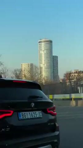 The video captures a stationary black BMW X3 in an urban setting. The vehicle's rear is visible, including its license plate "BG 1625-JC." In the background, tall, modern buildings stand against a clear blue sky. A traffic light with a lit green signal is visible to the left of the car. As the camera pans slightly, a large advertising billboard comes into view, promoting Lidl with the slogan "Niske cene svaki dan," meaning "Low prices every day." The overall environment suggests a city landscape with a mix of modern architecture and commercial advertising.