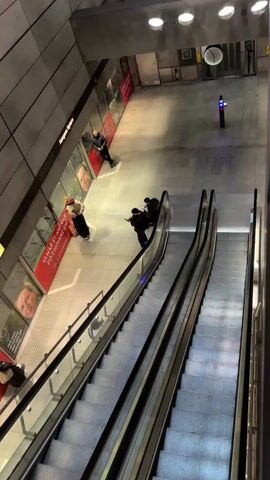 The camera looks downward into a modern subway station featuring two parallel escalators descending into the scene.  The station floor is tiled, and several people are visible. One individual stands near the left wall, looking at their phone. Another pair stands near the wall to the left closer to the camera. Two more individuals are riding the right escalator. There is a clock on the wall in the background. There are several advertisements along the left wall, behind the people.