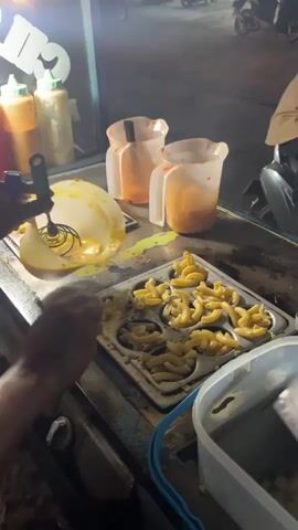 A person's hands are visible, preparing food at a food cart at night. The person is using a whisk to pour a creamy yellow substance, likely cheese sauce, from a bowl into a metal tray filled with macaroni pasta. The tray has individual circular molds, and the pasta is arranged within these molds. In the background, there are plastic jugs with red liquid, suggesting sauces or condiments. The environment appears to be a street scene at night, with lights from the cart and passing vehicles visible. The overall impression is of street food preparation.