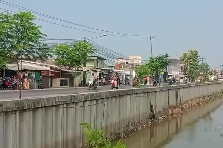 A long shot shows a street lined with shops and trees next to a concrete wall bordering a canal. Several people are riding motorcycles on the street, and various motorcycles are parked on the side. The canal contains murky water and some green vegetation. The sky is clear and bright, suggesting a sunny day.