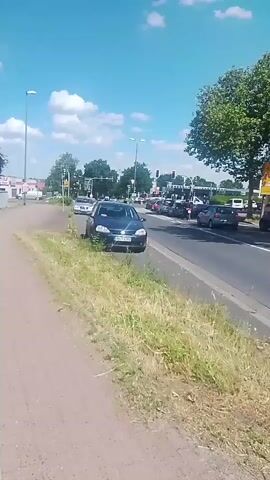 This is a short video clip showing a street scene in Bergkamen, Germany, on a sunny day with scattered clouds. The foreground is dominated by a patch of dry grass and a dirt path, suggesting an unpaved shoulder to a road. A dark blue hatchback car is parked on the grassy shoulder, partially obscuring the view of the road behind it. This car appears to be a Volkswagen Golf, with a license plate that reads "UN-T 4592".

The main road in the middle ground shows several other cars, some appearing to be stopped at a traffic light, while others are in motion. The traffic appears moderate. In the background, there are more cars, buildings that resemble shops or businesses, streetlights, and trees. A large, leafy green tree is prominent on the right side of the frame. The overall impression is a typical suburban or semi-urban environment during daylight hours. The weather appears warm and clear. There are no people clearly visible in the clip, and the focus is on the cars and the road environment. The camera is positioned low and slightly angled, giving a sense of being at ground level.
