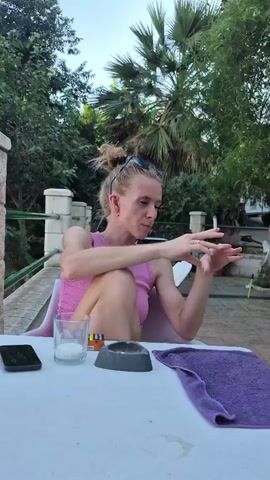 A woman sits at a white table outdoors, holding a cigarette between her fingers. She is wearing a pink sleeveless top and sunglasses on her head. The table has a cell phone, a glass with a white substance, an ashtray, and a purple towel. Behind her, lush green trees and palm trees create a tropical atmosphere. The scene appears to be set during daylight, likely late afternoon or early evening, given the warm lighting and the sky. The woman's expression suggests she is engaged in conversation, possibly contemplating the topic at hand. The setting suggests a relaxed, possibly vacation-like environment in Almáchar, Spain, with the greenery and outdoor seating indicative of a patio or garden area.