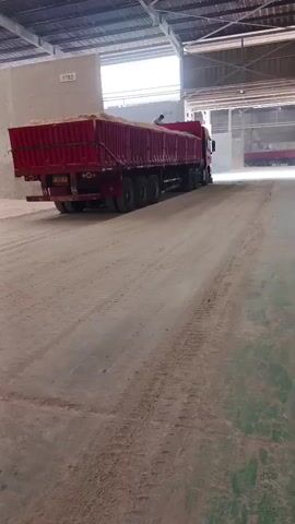 The video shows a red truck with a tarp-covered trailer full of light-colored material, likely grain or sand, driving into a large, open-sided industrial building. The floor of the building is covered in dust and tire tracks. The truck is moving from left to right across the frame, and the camera is positioned at a slight downward angle. The building has a metal roof supported by beams, and natural light is entering from the open sides and possibly openings in the roof. In the distance, another red truck can be seen inside the building. The overall impression is of an active industrial or agricultural facility.