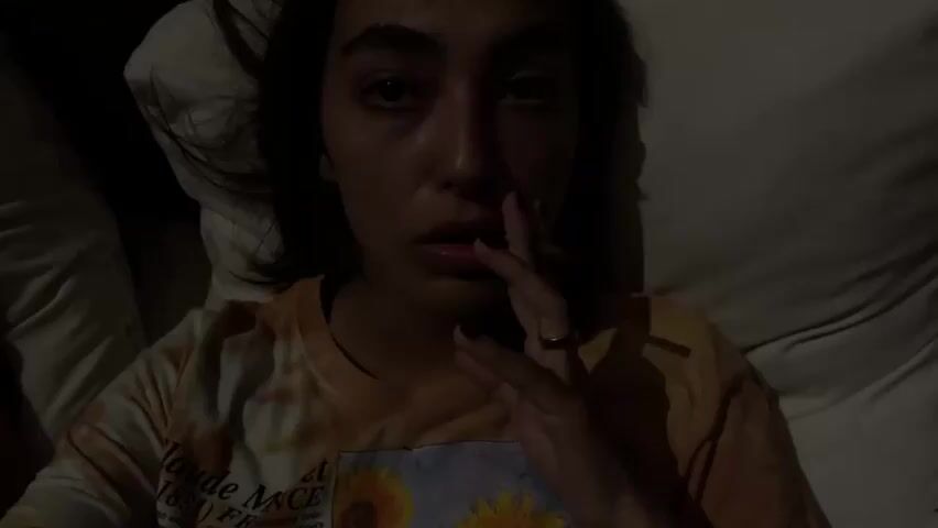 In dim lighting, a woman lies on a bed smoking what appears to be a hand-rolled cigarette. She is wearing a tie-dye t-shirt and has dark hair. A wedding ring is visible on her left hand. She is then seen turning her head to the right, where another woman lies, partially visible, in the same bed. The second woman has dark hair and is wearing a red and white striped top. Loud music plays in the background.