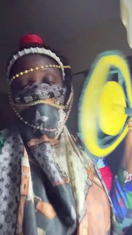 The video shows a person indoors, heavily covered in clothing and accessories, possibly for concealment. They wear a headpiece with beads and a red object on top, a patterned scarf covering most of their face, and a scarf or vest with various patterns. The person is holding a yellow plastic disc, which they briefly place on their head. The room appears dimly lit with minimal background details visible, including a wall and a curtain.