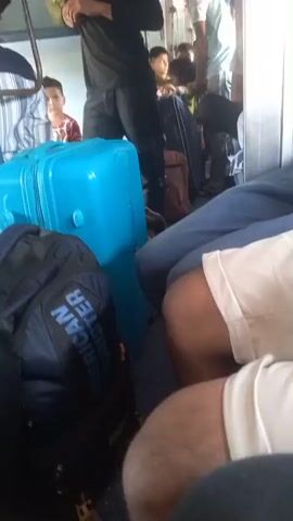 The video shows the interior of a train car.  Several people are standing or sitting, including a man wearing a dark blue polo shirt and a baseball cap, who is seated and looking down at his phone.  Luggage, including a blue suitcase and several backpacks, is visible in the aisle and between the seats.  Other passengers are milling about or sitting. The train appears crowded. The camera moves erratically at times.