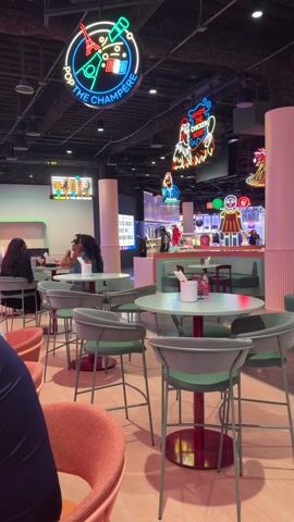 The video shows the interior of a brightly decorated restaurant, possibly within a larger shopping complex. Several round tables with pastel green chairs and red bases are prominent in the foreground, with some patrons seated at them. Further into the space are upholstered booths in a similar pastel green. Walls are tiled in shades of pink and peach, and various neon signs hang from the ceiling, advertising different food or drink items. A sign for "Netflix Bites" is visible, suggesting a themed dining experience. People are seated at tables in the distance, eating. The overall atmosphere is vibrant and modern.