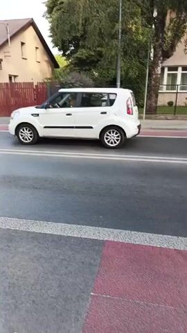 A white Kia Soul drives down a street. The car is moving from left to right in the frame. The driver is visible inside the car, as is at least one passenger. The car is driving on a road with white lane markings and red and white crosswalk markings. On the left side of the road is a wooden fence and a house. On the right side of the road are more buildings and some trees. The sky is visible at the top of the frame. The video is shot from a low angle.