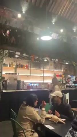 The video shows the interior of a busy bar or restaurant. Several groups of people are seated at tables, eating and talking. A bar runs along the back of the room, attended by two bartenders. The space is lit by overhead lights and decorated with greenery. There is a disco ball hanging from the ceiling. The room has a high ceiling with visible wooden beams.