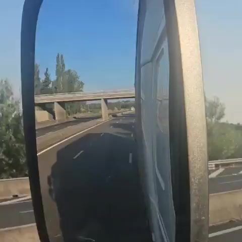 The video captures a view through a side mirror of a vehicle traveling on a highway. The mirror reflects a highway with multiple lanes, a concrete barrier, and a red car visible in the distance. A pedestrian bridge spans across the highway. Trees and vegetation line the sides of the road. The sky is clear and blue, with a hazy or smoky cloud formation visible beyond the bridge. The camera perspective suggests the vehicle is a truck, given the size and angle of the mirror.